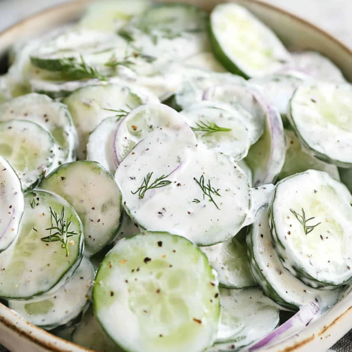 Close-up of cucumber salad with creamy dressing, garnished with dill sprigs on a checkered cloth.