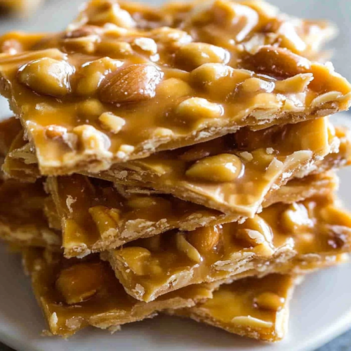 Close-up of crunchy caramel candy with embedded peanuts, broken into thin shards and served on a white plate.