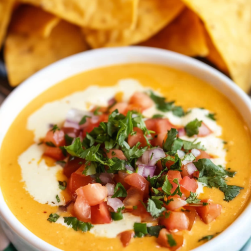 Close-up of smooth cheese dip with a drizzle of sour cream and colorful garnishes.