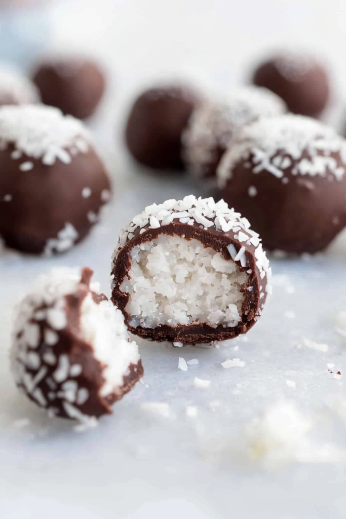 Close-up of coconut truffles coated in chocolate and topped with shredded coconut on a white surface.