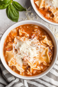 Bowl of tomato-based soup with lasagna noodles, ground meat, and fresh basil on top.