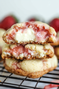 Soft cookies with a creamy cheesecake center and visible strawberry filling, stacked on a cooling rack