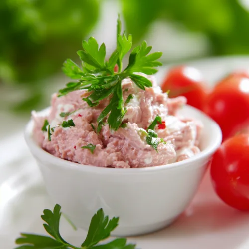 Close-up of a creamy meat salad served with crackers and leafy greens in the background.
