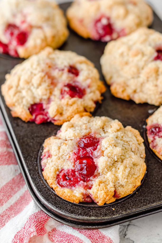 Golden muffins with juicy cherry pieces peeking through the crumbly tops in a rustic tin.