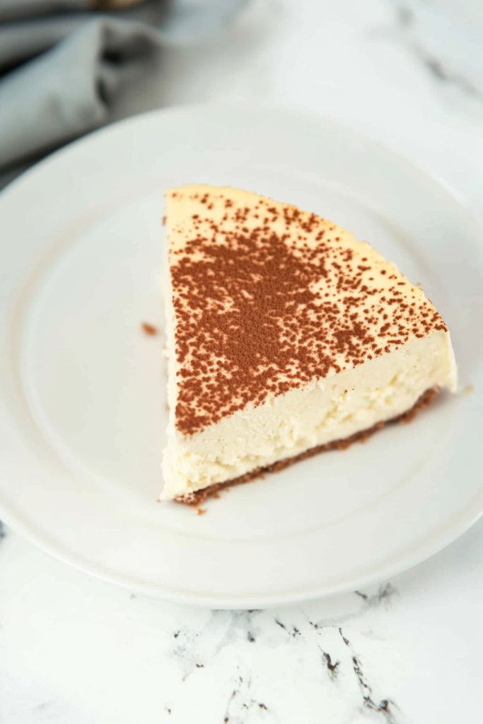 Slice of creamy baked cheesecake with a graham cracker crust and cocoa dusting on top.