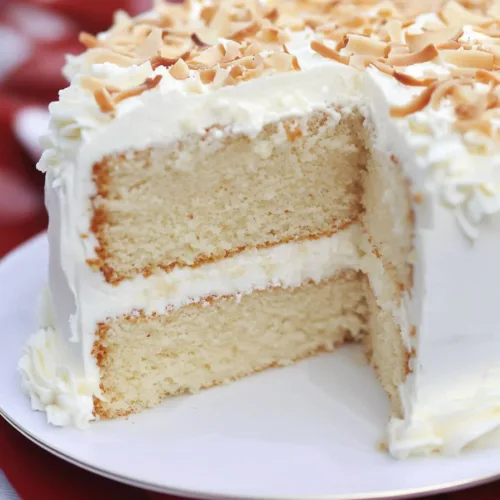 A frosted two-layer cake topped with toasted coconut flakes, presented on a white plate with a festive red cloth beneath.