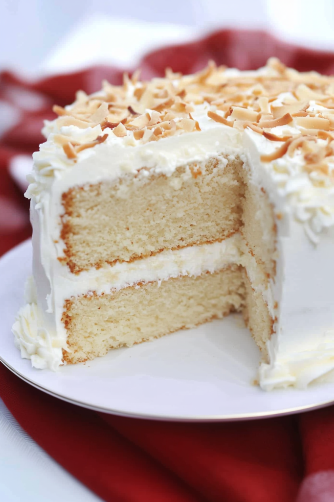 A frosted two-layer cake topped with toasted coconut flakes, presented on a white plate with a festive red cloth beneath.
