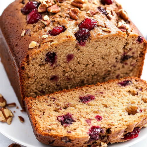 Thick slice of tender cake showing bursts of red cranberries and a nutty pecan topping.
