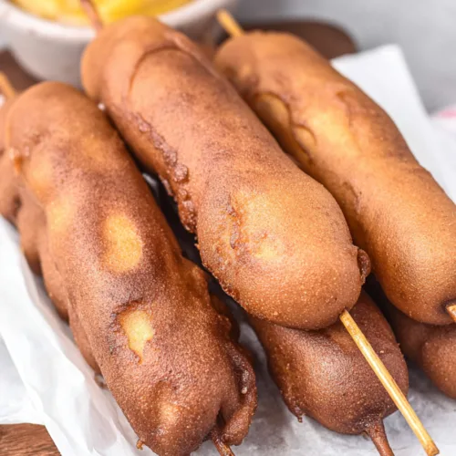 Freshly fried corn dogs showing crunchy exterior, plated simply with parchment paper and mustard on the side.
