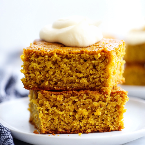 Moist pumpkin cornbread squares stacked on a white plate with a dollop of whipped cream on top.