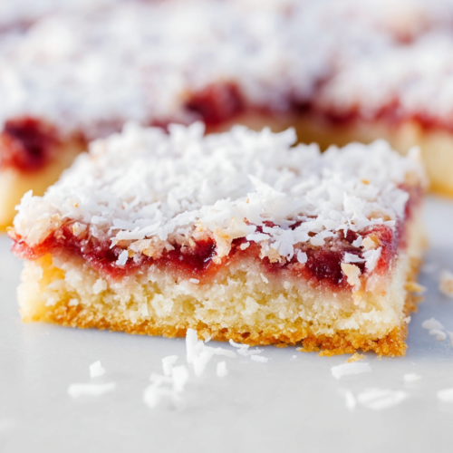 Close-up of layered bars showing red jam center and toasted coconut flakes on top.