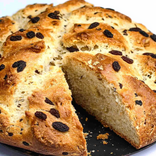 Round loaf of golden-brown bread on a dark plate, with raisins visible on the crust and a slice cut to show the airy interior.
