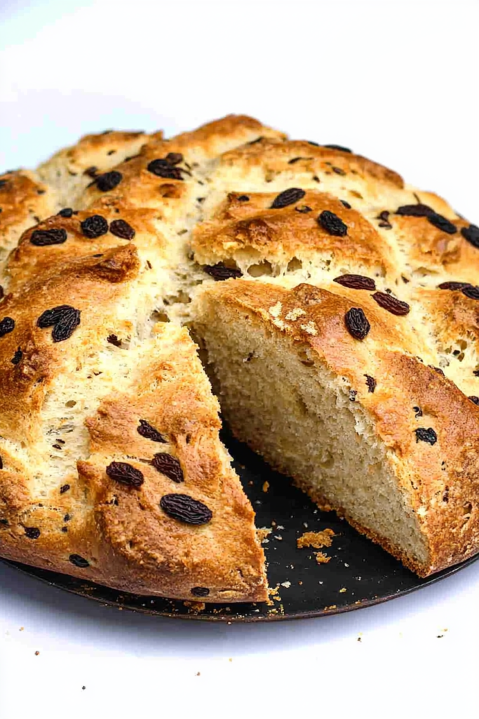 Round loaf of golden-brown bread on a dark plate, with raisins visible on the crust and a slice cut to show the airy interior.