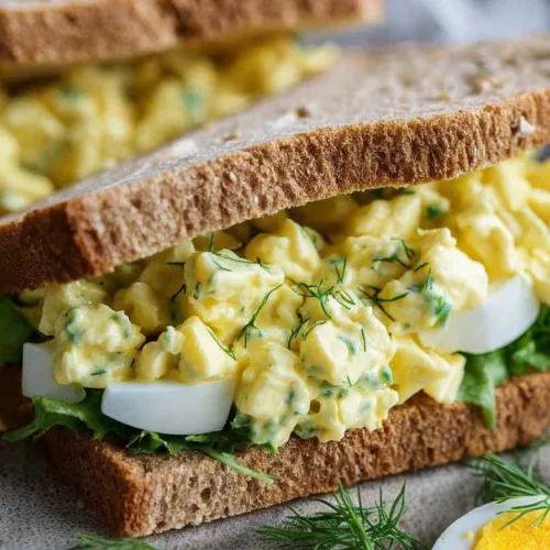 Whole grain sandwich filled with creamy egg salad and fresh herbs, garnished with dill sprigs.