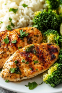 Close-up of juicy baked chicken breast garnished with herbs, paired with rice and fresh green broccoli.
