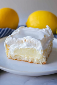 Creamy no-bake lemon cake slice with a crumb crust and fresh lemons in the background.