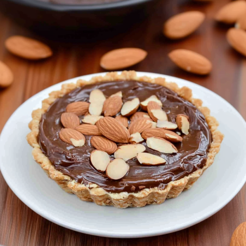 Homemade tart with smooth chocolate surface and nutty topping, presented on a wooden table.