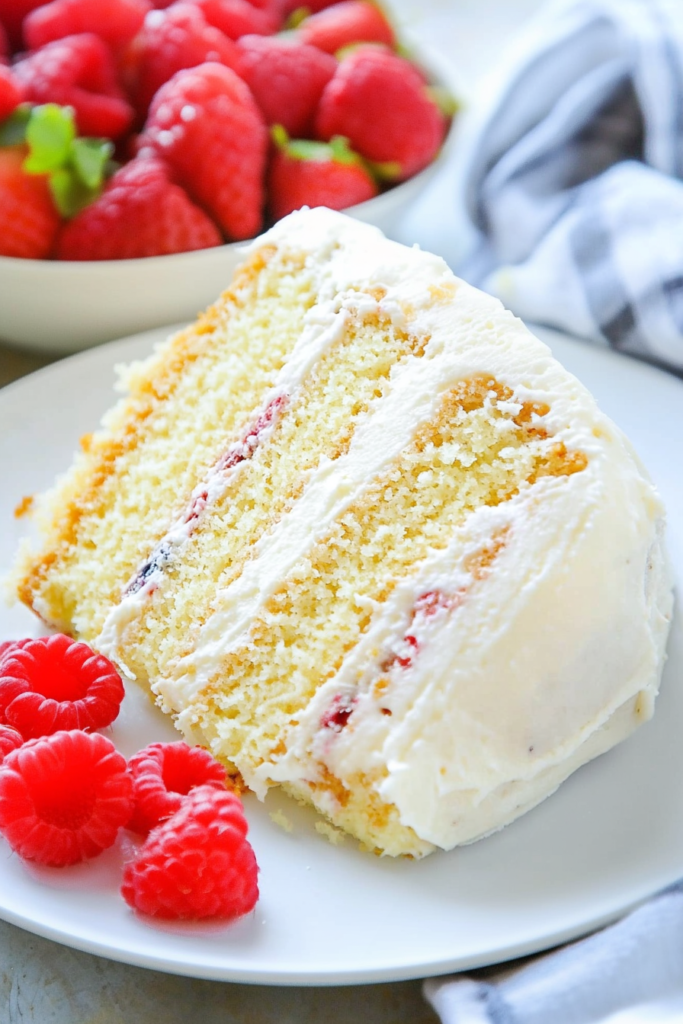 โSlice of vanilla sponge cake with raspberry filling and cream frosting, served with fresh berries