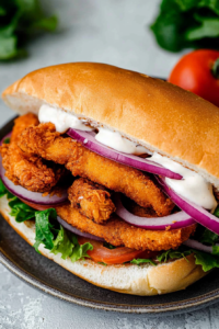 Golden-brown fried seafood tucked into a sandwich bun with fresh vegetables and creamy sauce.