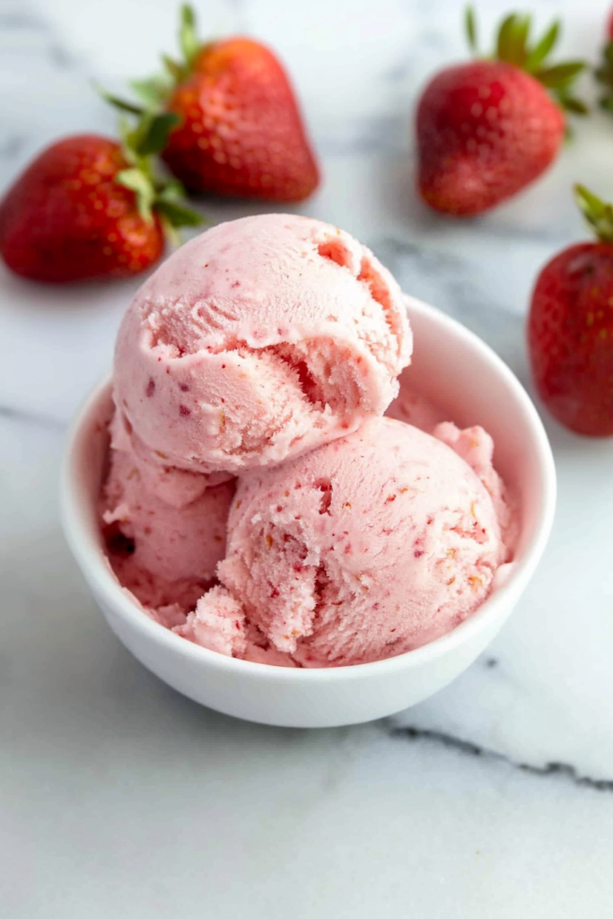 Bowl of frozen dessert with strawberries in the background on a marble surface.