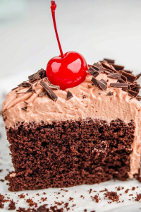 Moist chocolate cake slice topped with frosting, chocolate shavings, and a bright cherry.
