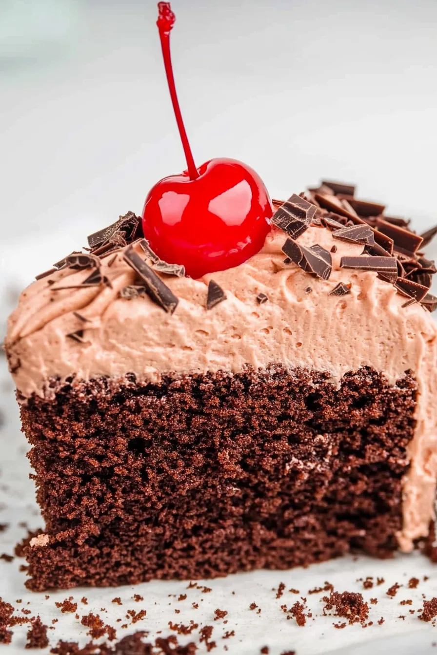 Moist chocolate cake slice topped with frosting, chocolate shavings, and a bright cherry.