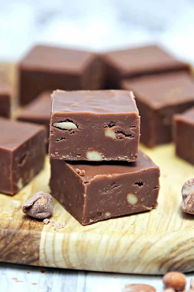 Stacked squares of glossy chocolate fudge with nuts on a wooden board.