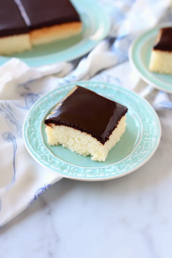 Slice of golden sponge cake topped with glossy chocolate frosting on a pastel plate.