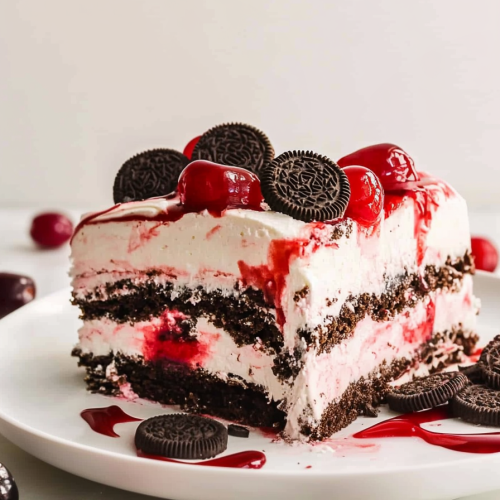 Cherryโtopped cake slice surrounded by syrup and scattered cookie crumbs on a white plate.