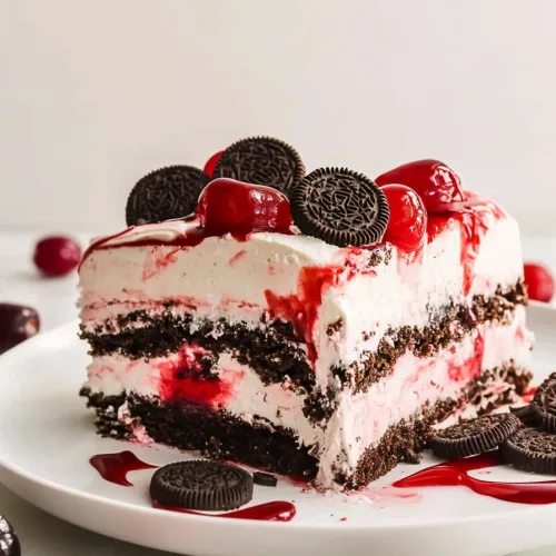 Cherryโtopped cake slice surrounded by syrup and scattered cookie crumbs on a white plate.