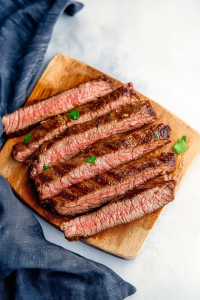 Slices of juicy grilled steak with visible char marks, arranged neatly on a wooden cutting board beside a creamy dipping sauce.