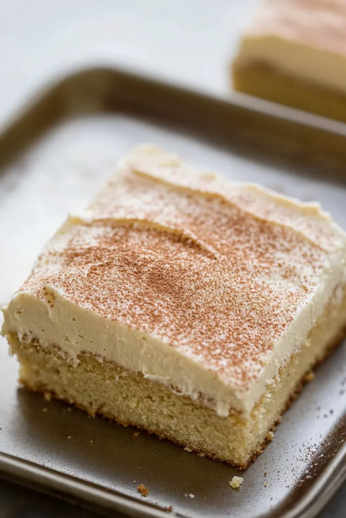 Large sheet cake slice on a plate, showing moist yellow layers topped with creamy frosting and a dusting of spice.