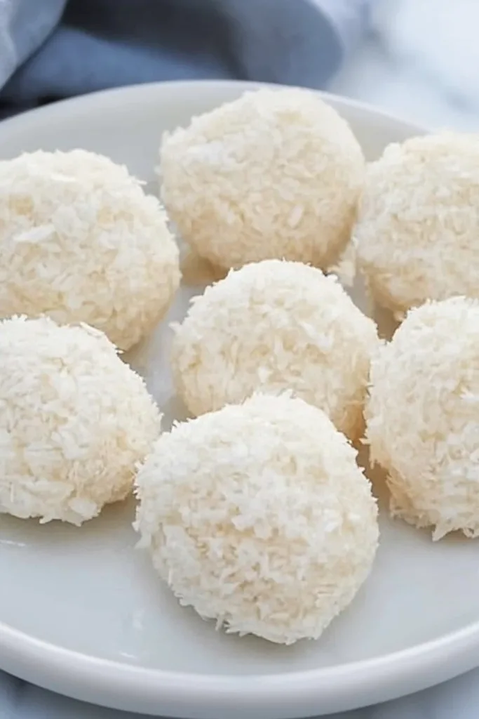 Round coconut-covered treats on marble, with one showing a creamy bite inside.