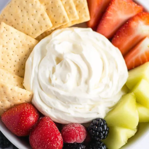 A bowl of creamy dip surrounded by fresh strawberries, melon, and berries with crackers for dipping