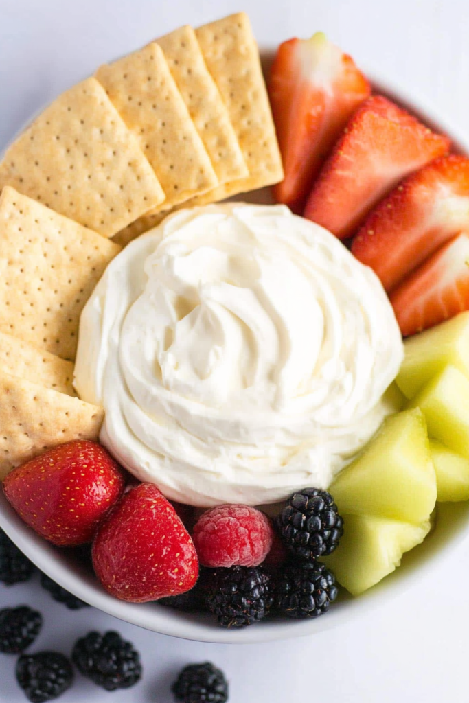 A bowl of creamy dip surrounded by fresh strawberries, melon, and berries with crackers for dipping