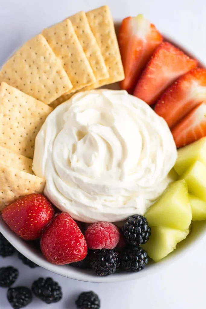 A bowl of creamy dip surrounded by fresh strawberries, melon, and berries with crackers for dipping