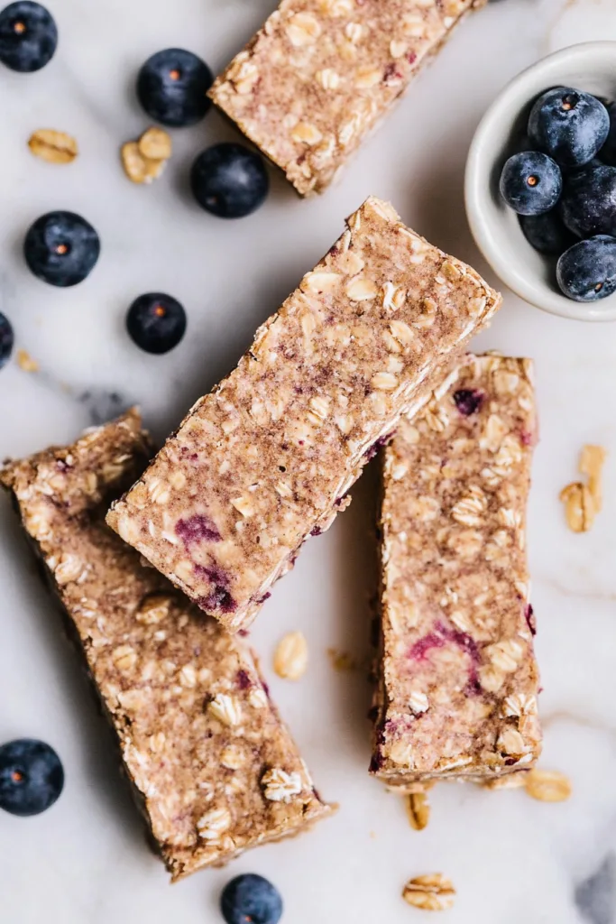 Homemade oat bars with blueberries scattered around on a light surface.