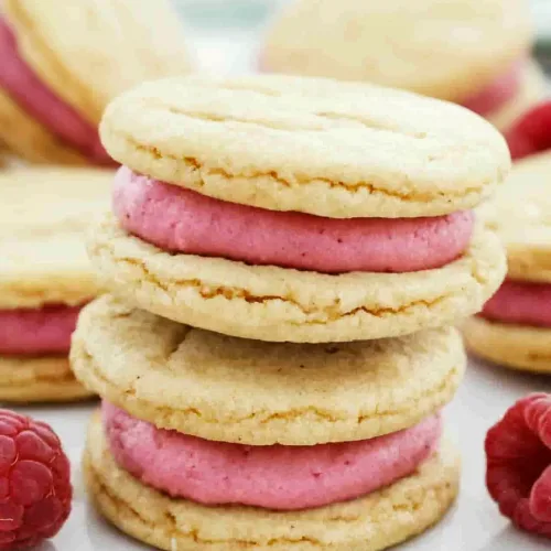 A plate of pastel cookies with creamy raspberry centers, styled with scattered berries for contrast.