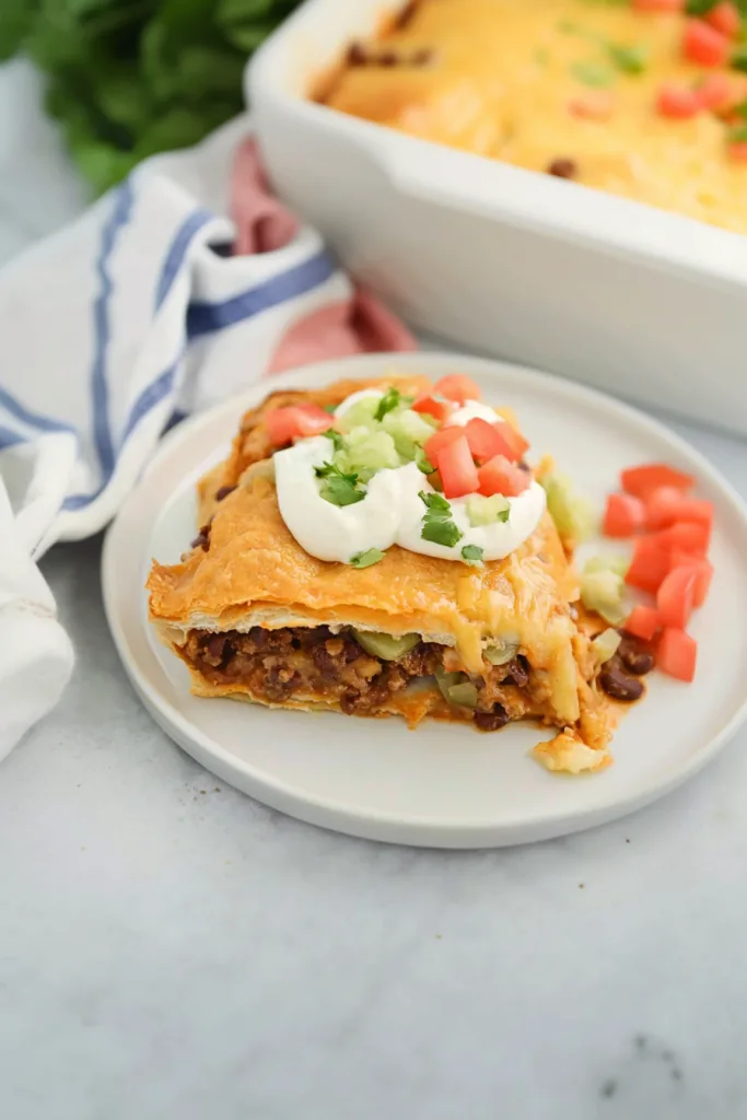 Slice of cheesy taco casserole topped with lettuce, tomatoes, and a dollop of sour cream.