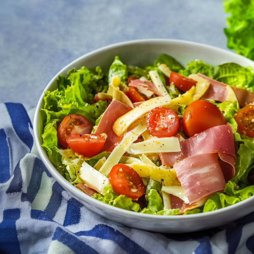 Fresh lettuce tossed with ham, cheese strips, and cherry tomatoes in a white bowl.