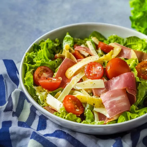 Fresh lettuce tossed with ham, cheese strips, and cherry tomatoes in a white bowl.