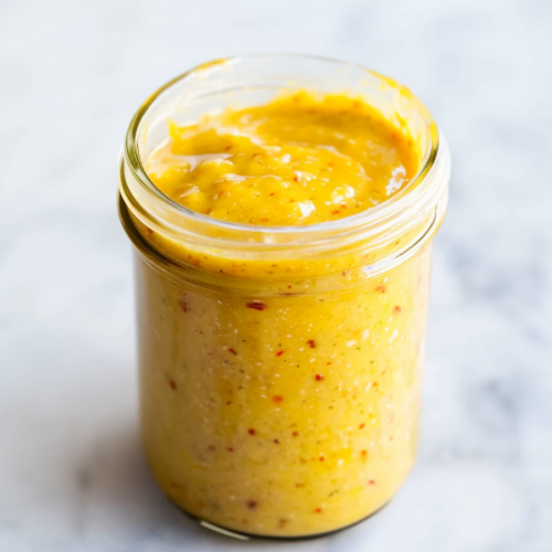 Glass jar of thick yellow mustard with red and black specks throughout.