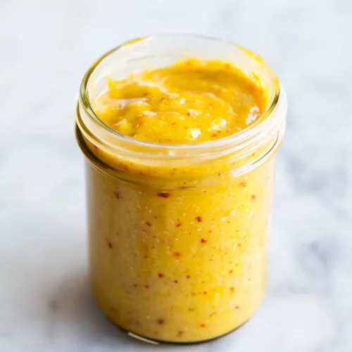 Glass jar of thick yellow mustard with red and black specks throughout.