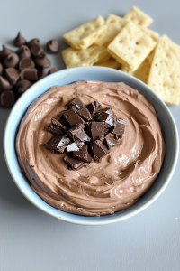 Bowl of creamy chocolate dip surrounded by crackers and chocolate chips.