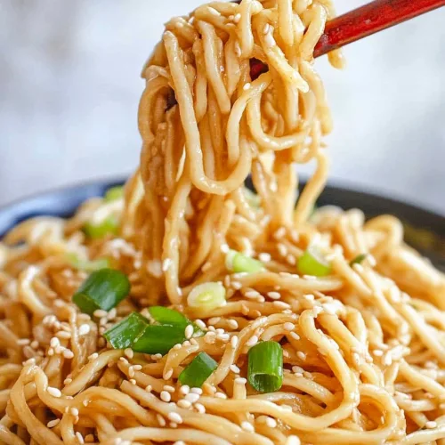 Glossy noodles lifted with red chopsticks, sprinkled with sesame seeds and green onions.