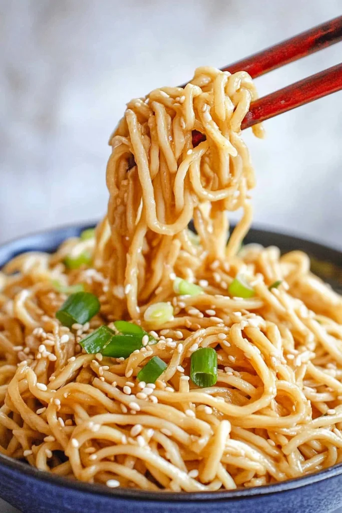 Glossy noodles lifted with red chopsticks, sprinkled with sesame seeds and green onions.
