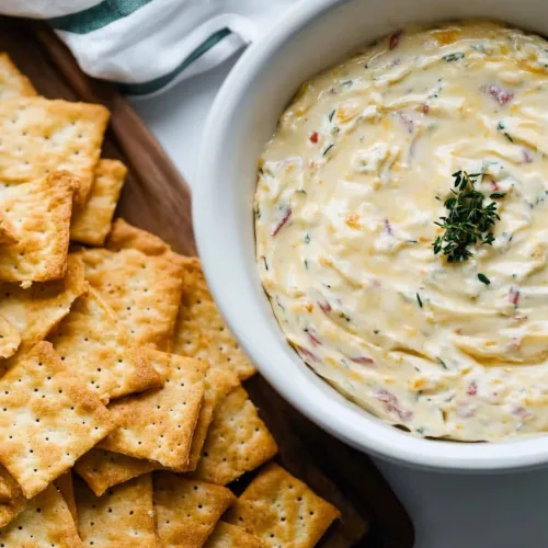 A creamy dip in a white bowl, topped with fresh herbs and surrounded by golden crackers.