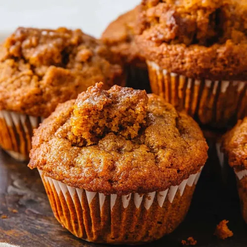 Freshly baked golden-brown muffins in white paper liners, resting on a wooden surface with scattered crumbs.