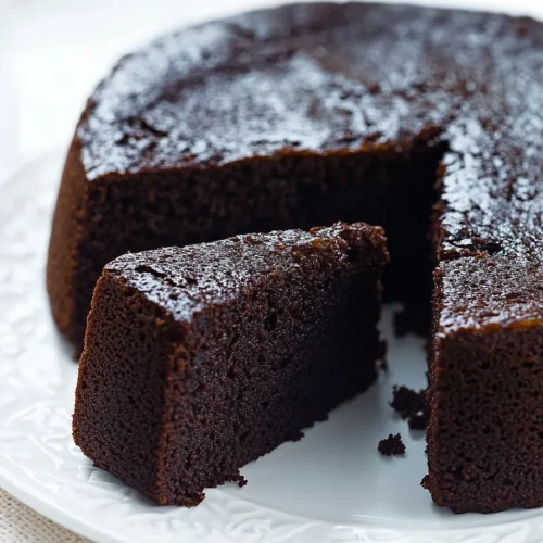 Rich, dark chocolate-colored cake with a slice served on a white plate.
