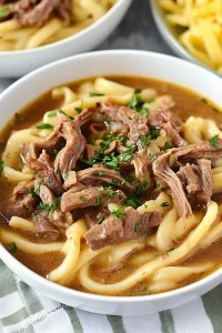 Bowl of tender noodles in rich brown sauce with beef slices on top.